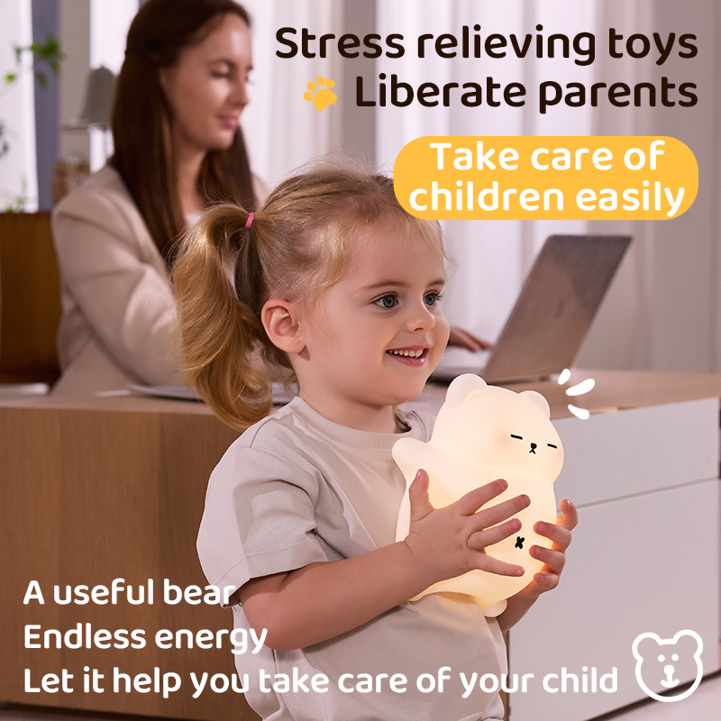 Smiling young girl happily holding a glowing, soft bear-shaped silicone night light while a woman works on a laptop in the background.